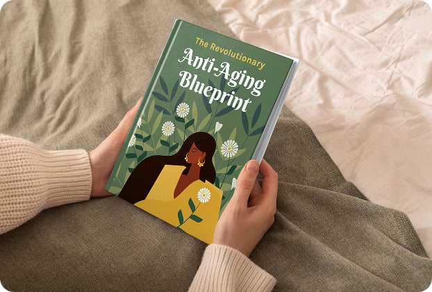 Person holding a book titled 'The Revolutionary Anti-Aging Blueprint' with an illustrated cover featuring a woman surrounded by green leaves and white flowers, while sitting on a cozy bed with a blanket.